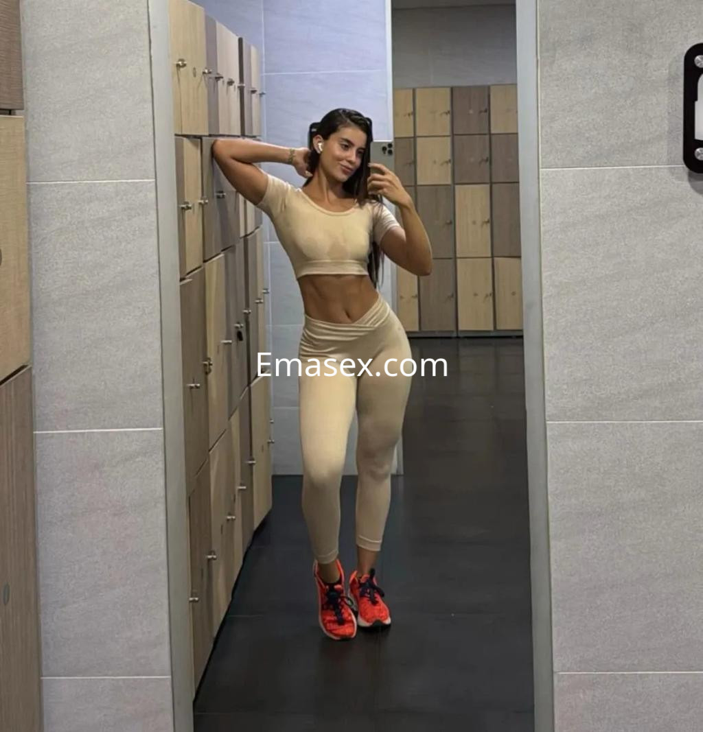 Puta en Sevilla posando en un gimnasio con un conjunto deportivo beige, sonriendo frente al espejo.