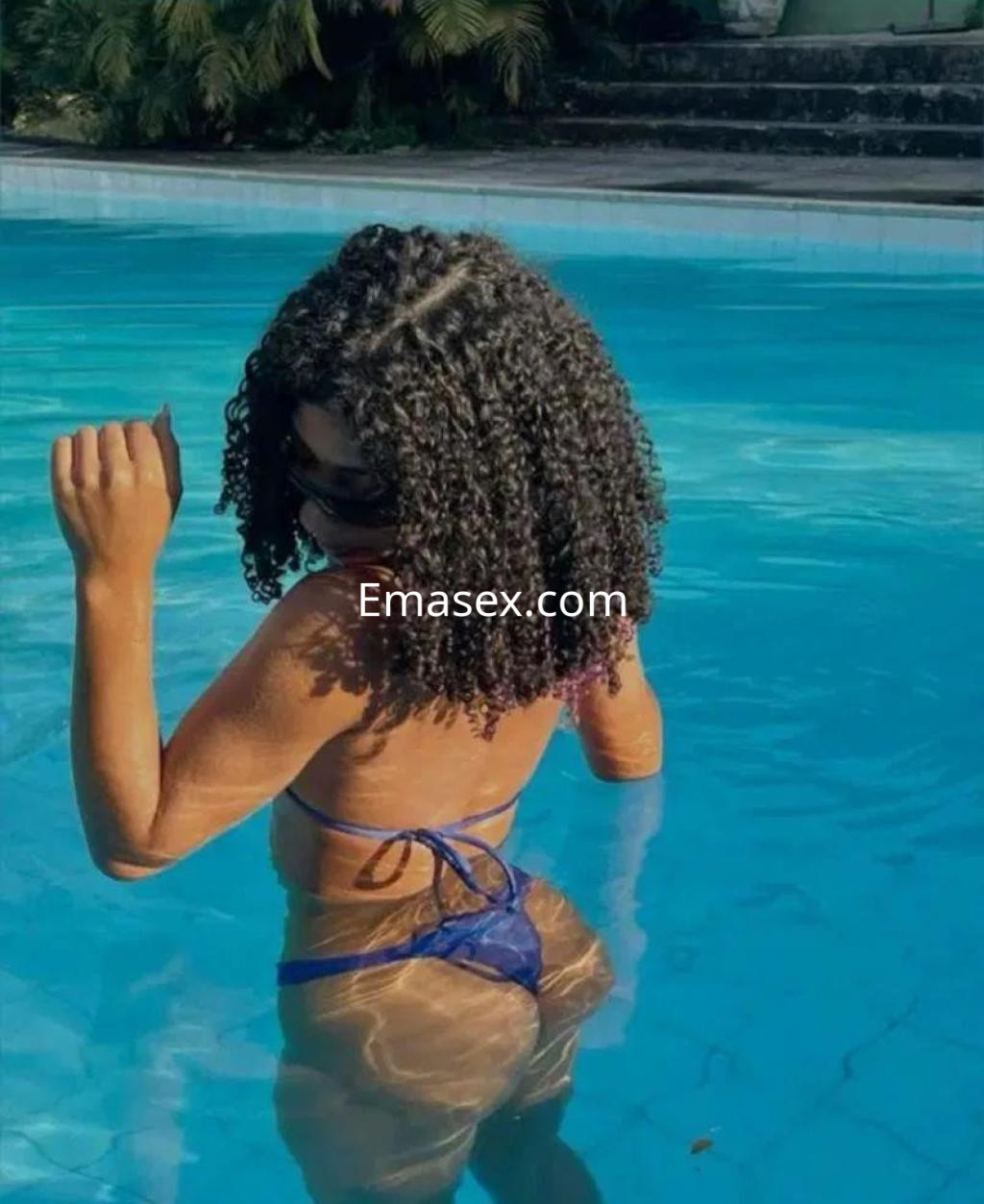 Escort en Sevilla, con cabello rizado y piel bronceada, posando de espaldas en una piscina.