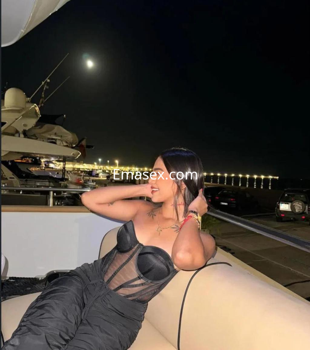 Escort en Sevilla, posando en un yate nocturno, viste un corset negro y pantalones oscuros, con el mar de fondo.