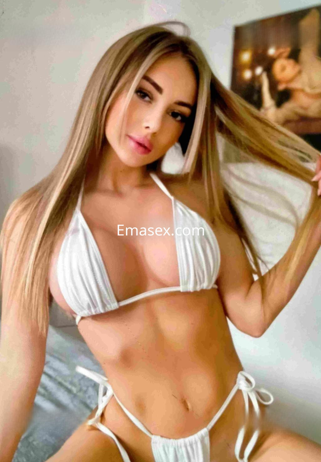 Escort en Barcelona con cabello largo y rubio, vestida con un bikini blanco, posando en un entorno interior.