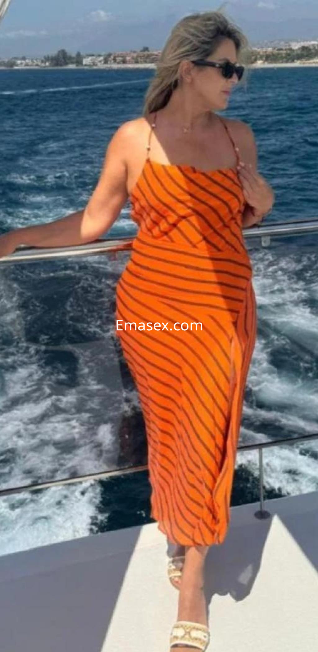 Escort en Coruña, luciendo un vestido largo naranja con rayas mientras posa en un yate junto al mar.