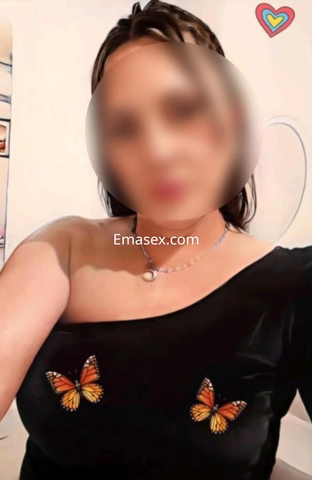 Escort en Huelva luciendo un top negro con mariposas, cabello oscuro y un ambiente acogedor de fondo.