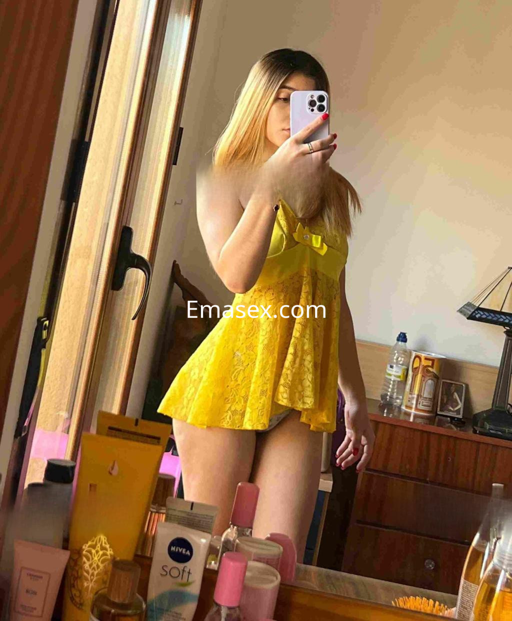 Escort en Toledo luciendo un vestido amarillo con encaje, posando frente a un espejo en un ambiente interior.