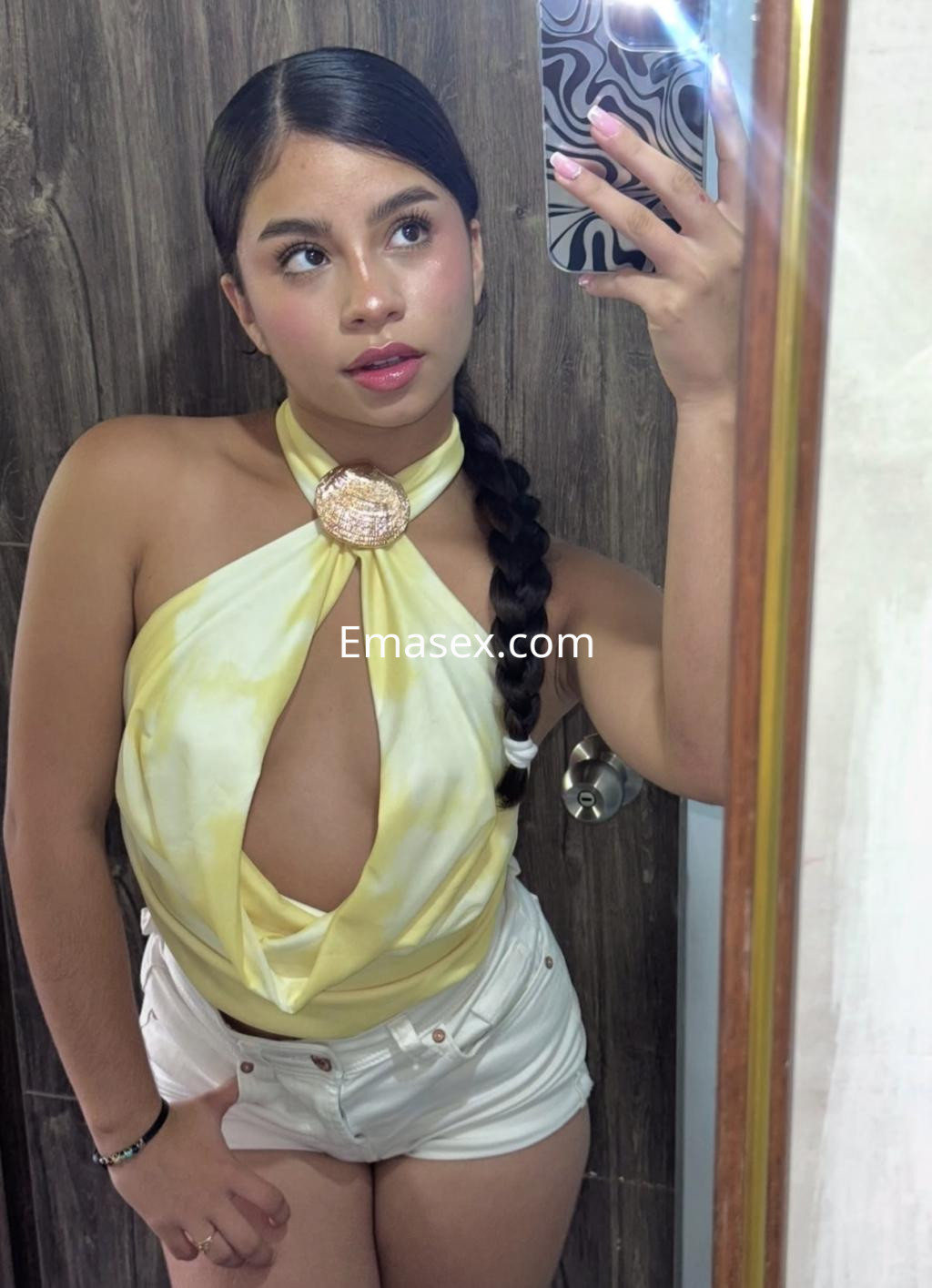 La escort en Bilbao lleva un top amarillo con un escote pronunciado y shorts blancos, posando frente a un espejo.