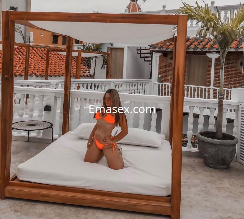 Escort en Irún, posando en una cama al aire libre con un bikini naranja, rodeada de un ambiente tropical.