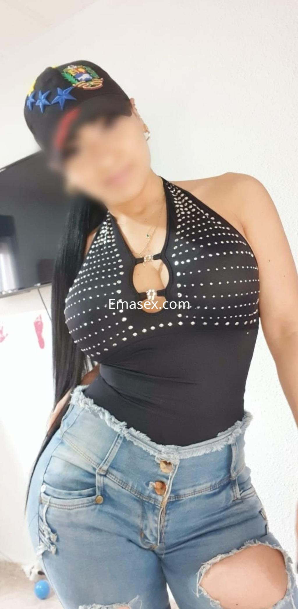 La puta viste un top negro con detalles brillantes y shorts de mezclilla, posando en un ambiente interior.