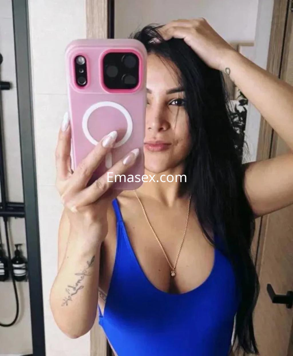 Escort en Albacete, con cabello largo y oscuro, luciendo un bañador azul frente al espejo en un baño moderno.