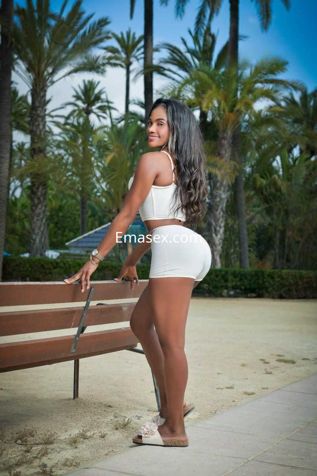 Puta con cabello largo y lacio, vestida con un conjunto blanco, posando de perfil en un parque de Alicante.