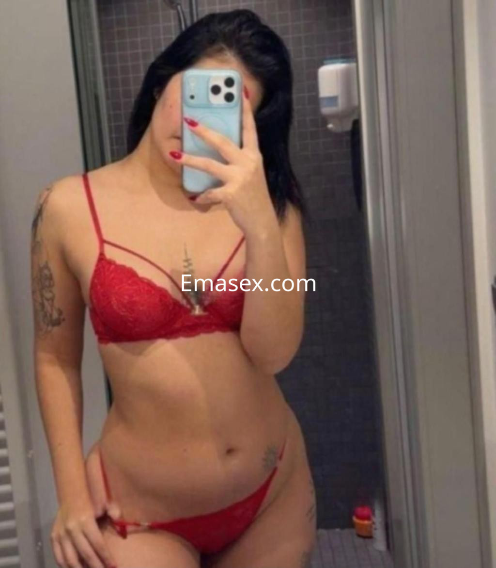 Escort en Badajoz con lencería roja, mostrando su figura en un espejo, con cabello oscuro y tatuajes visibles.
