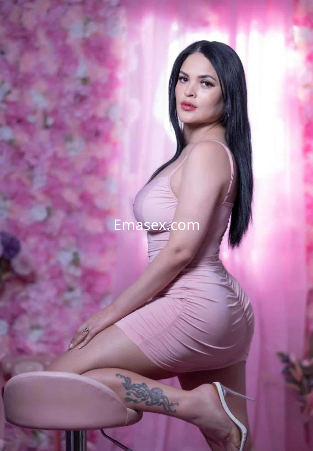 Escort en Barcelona con vestido rosa ajustado, posando sobre un taburete en un entorno decorado con flores.