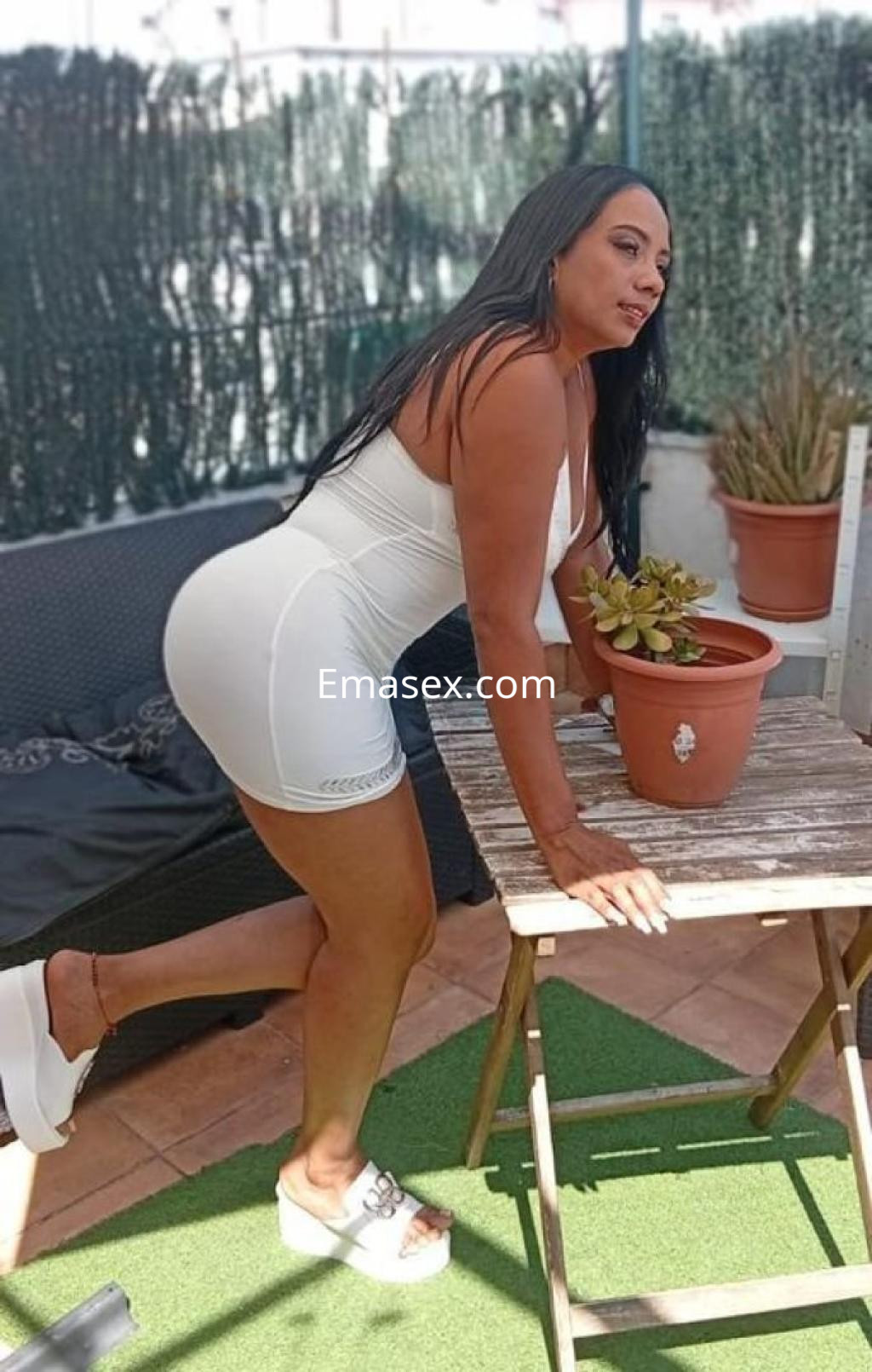 La puta posa en Barcelona, luciendo un vestido blanco ajustado y tacones, con una planta en la mesa.