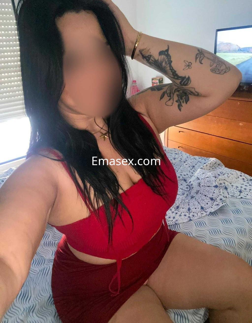 Escort en Burgos posando en una cama, con vestido rojo y tatuajes visibles, cabello largo y oscuro.