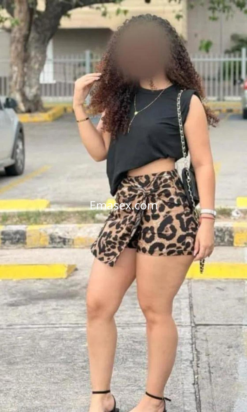 Escort en Cáceres con cabello rizado, viste un top negro y shorts de leopardo, posando en un entorno urbano.