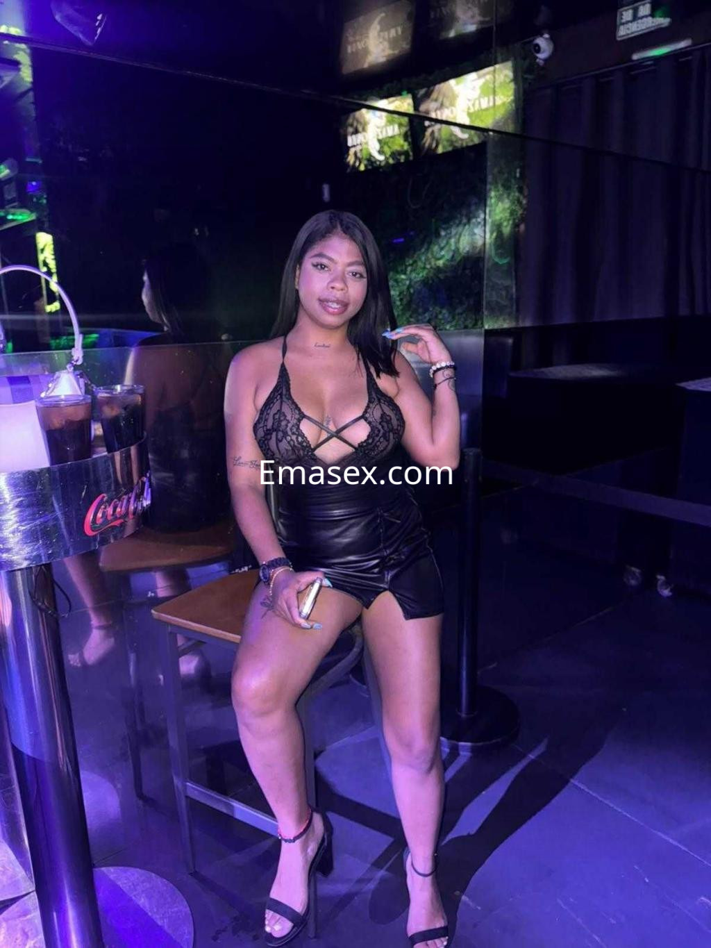 La escort en Castellón viste lencería negra y falda de cuero, posando con confianza en un bar iluminado.