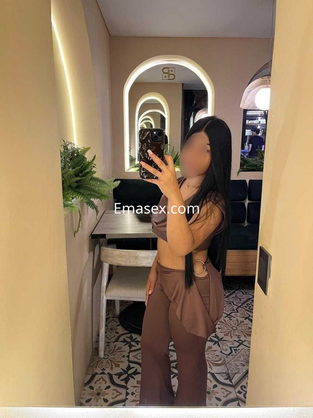 Escort con cabello largo y lacio, vistiendo un conjunto marrón, se toma una selfie en un ambiente elegante de Castellón.