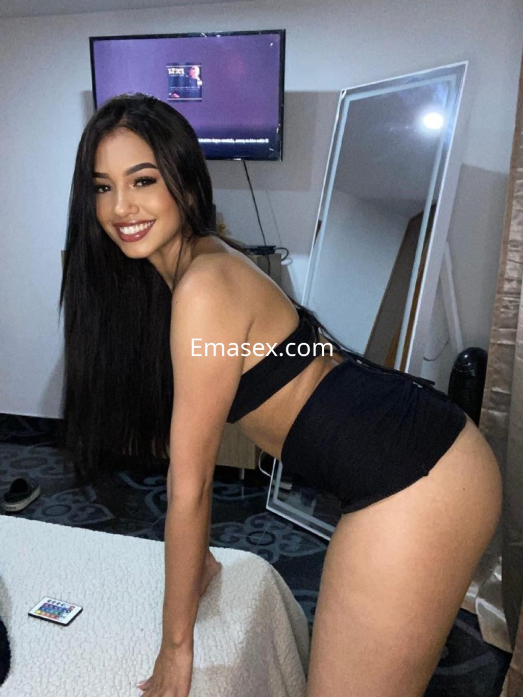 Escort en Granada, con cabello largo y oscuro, viste un top negro y shorts, sonriendo en un ambiente interior.