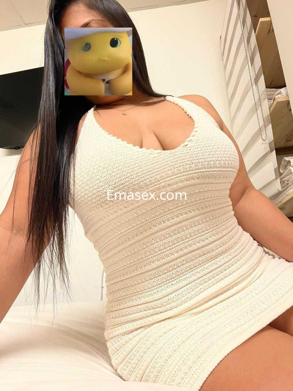 La puta posa en una habitación con un vestido blanco ajustado que resalta su figura, con cabello largo y lacio en Huelva.