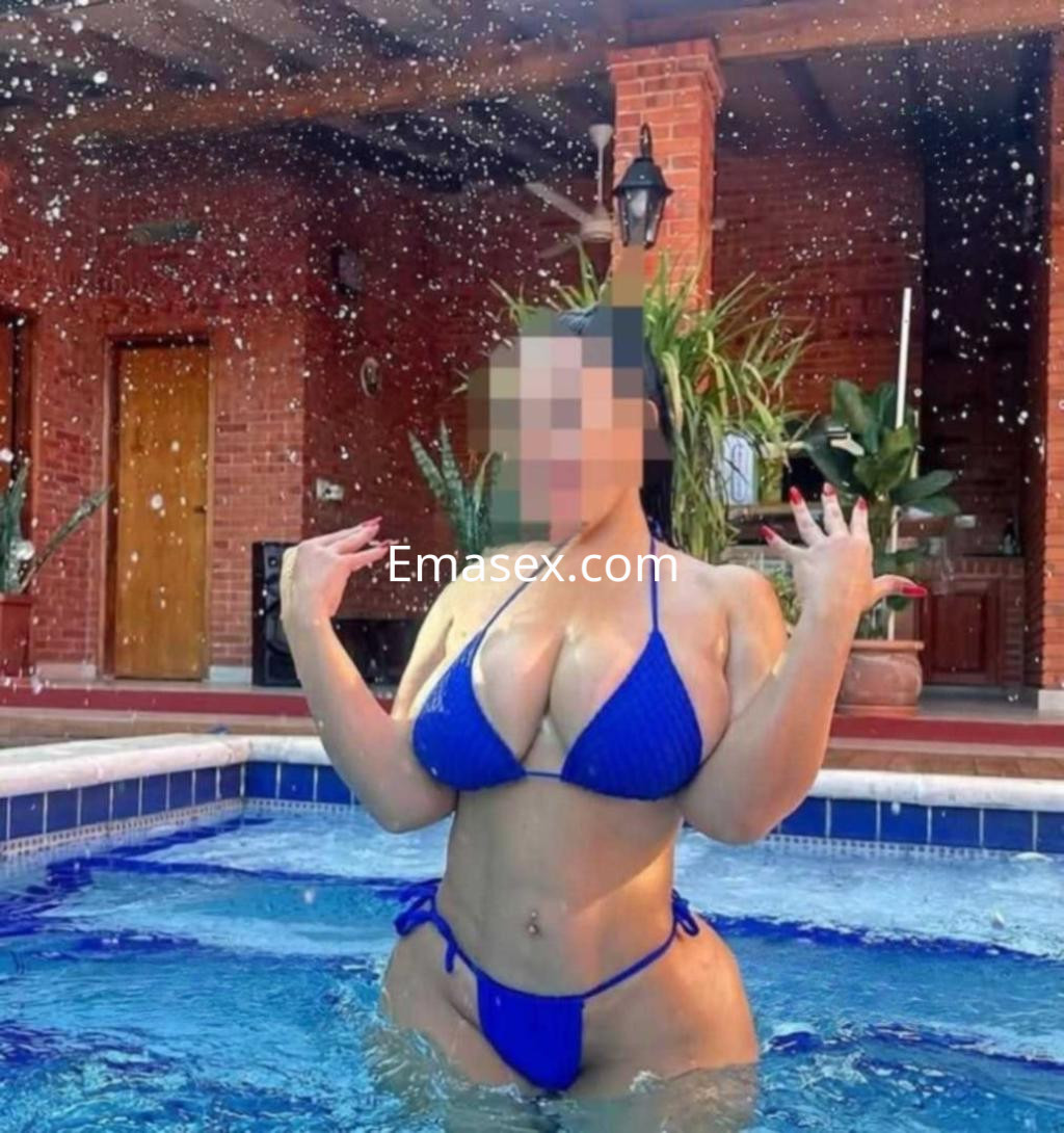 Puta en León posando en una piscina con un bikini azul, rodeada de agua y un ambiente tropical.
