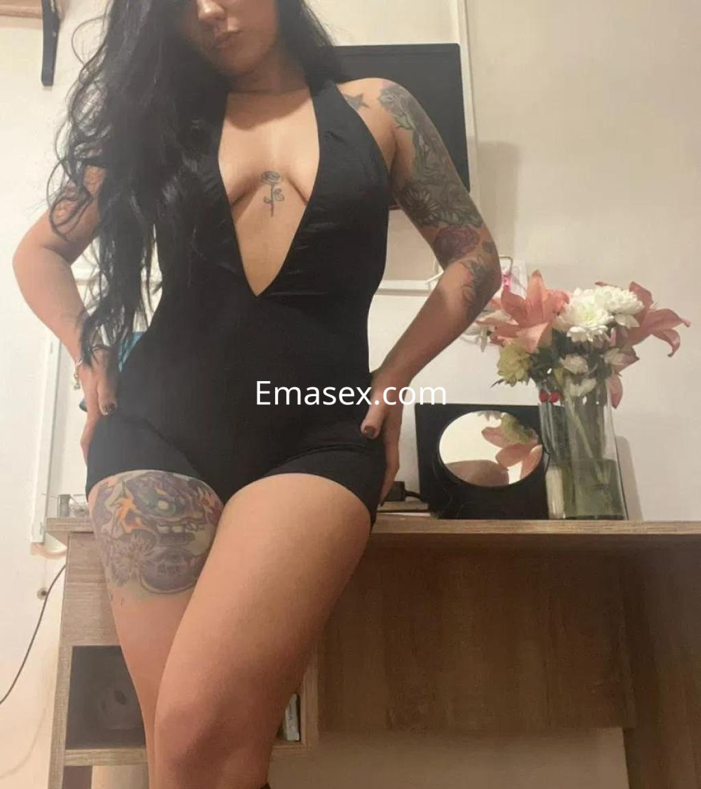 Escort en Lugo con un body negro de escote pronunciado, luciendo tatuajes en brazos y piernas, en un entorno acogedor.