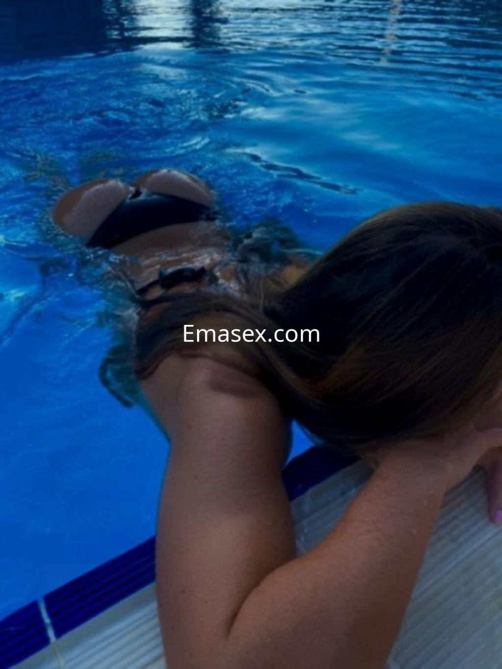 Escort en Lugo, sumergida en una piscina, con cabello castaño y traje de baño oscuro, posando de espaldas.