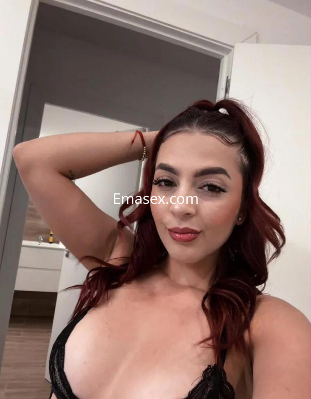 Escort en Madrid con cabello rizado y rojo, vistiendo lencería negra, posando con una mano en la cabeza.