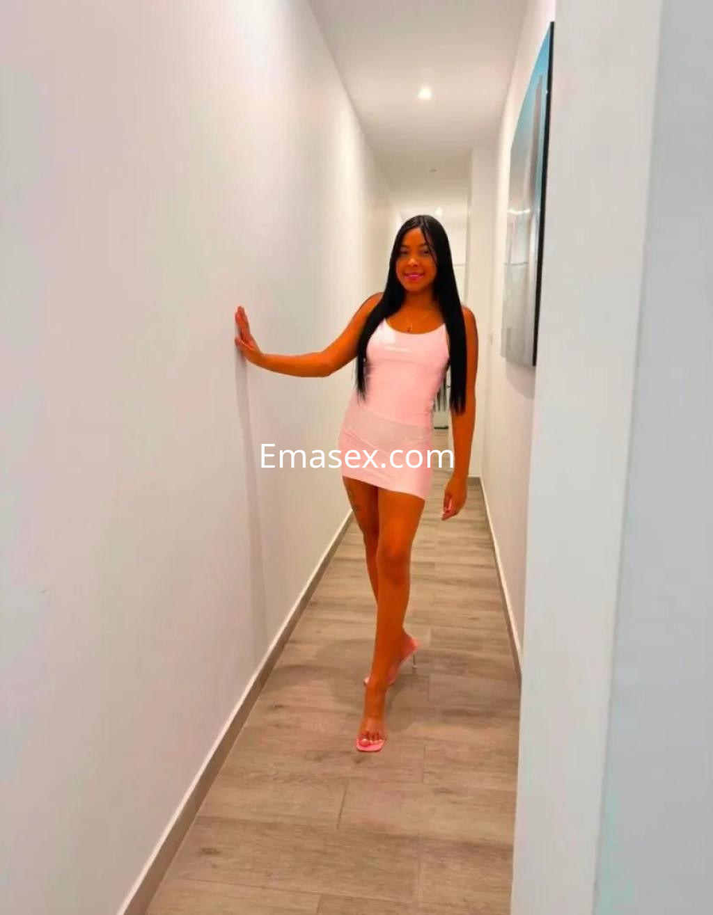 Puta sonriente camina por un pasillo con paredes blancas, luciendo un vestido corto rosa y sandalias.