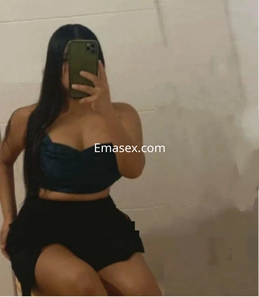 Puta en Sevilla con blusa de tirantes azul oscuro y falda corta, posando sentada y mirando al espejo.