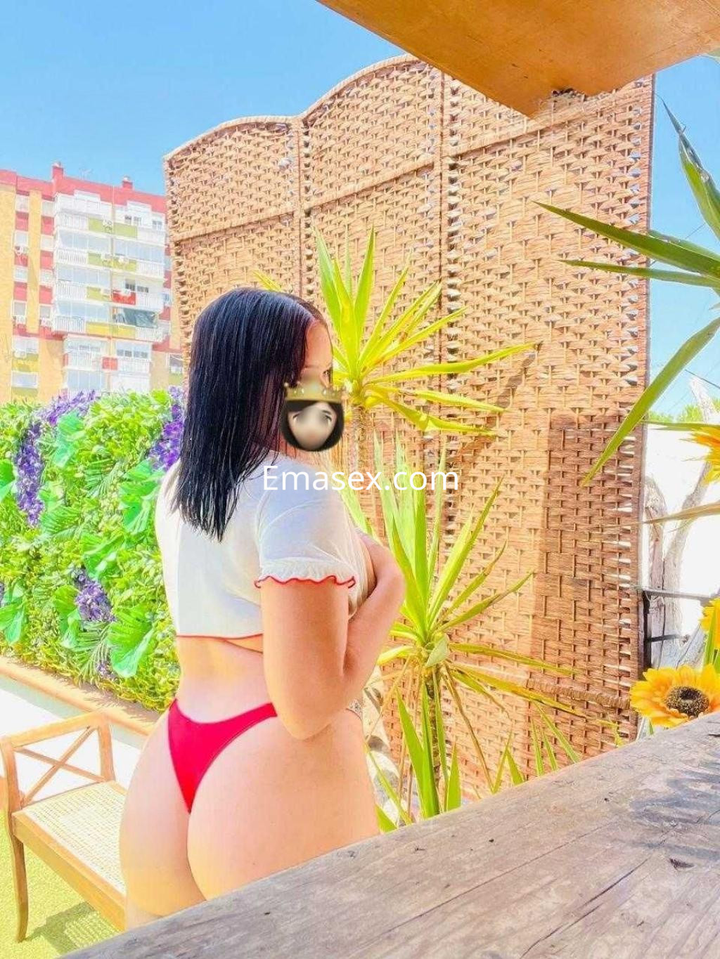 Escort en Sevilla con cabello oscuro, luciendo un top blanco y bikini rojo, de pie frente a un fondo de plantas.