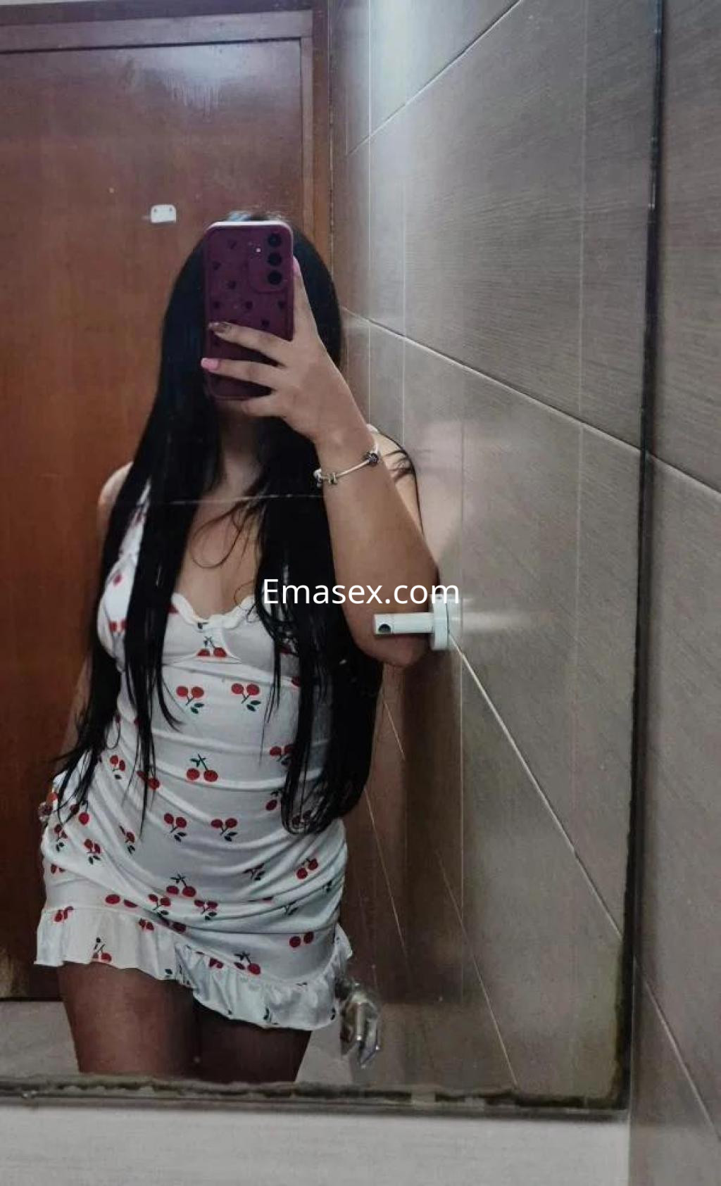 Escort en Sevilla posando en un espejo, viste un vestido blanco con estampado de cerezas y tiene el cabello largo.