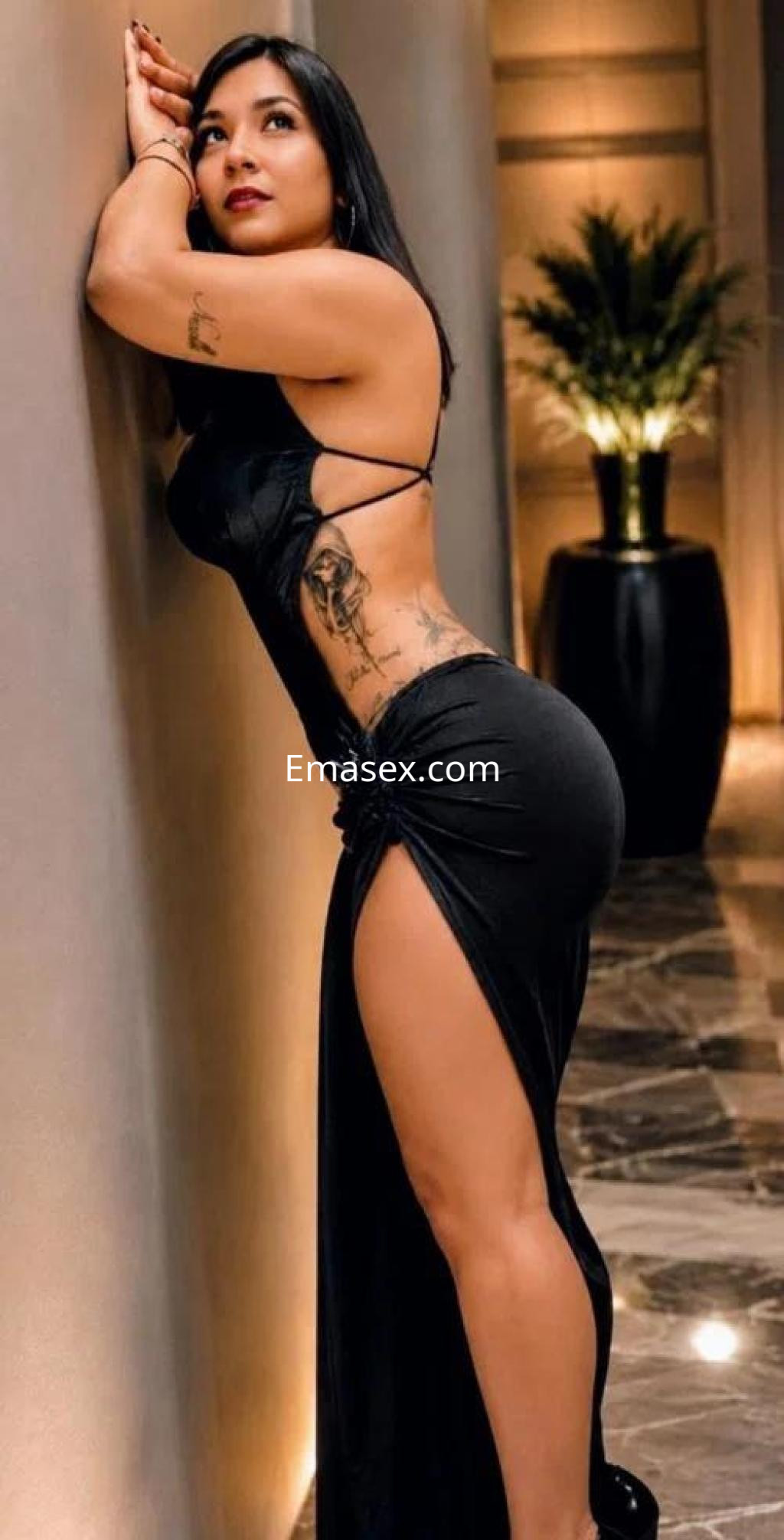 Escort en Tarragona posando de lado con un vestido negro ajustado y una abertura alta, mostrando un tatuaje en el brazo.
