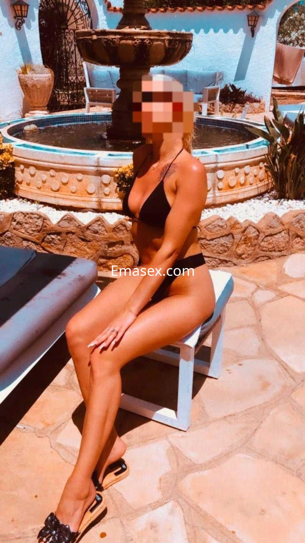Escort en Tarragona sentada junto a una fuente, luciendo un bikini negro y sandalias, con cabello corto y rubio.