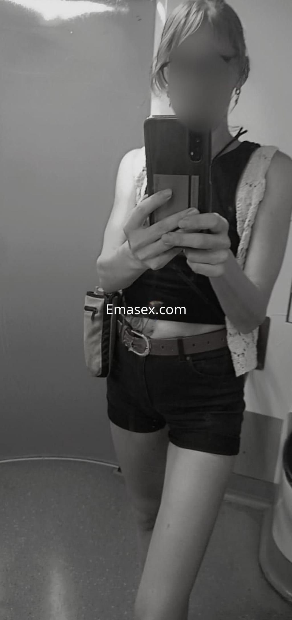 Puta en Tarragona se toma una selfie en un espejo, luciendo un top negro y shorts cortos, con cabello rubio.