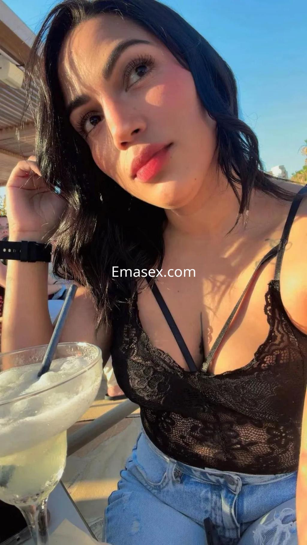 Escort en Toledo con cabello oscuro y lacio, luciendo un top de encaje negro y jeans, sosteniendo una bebida.