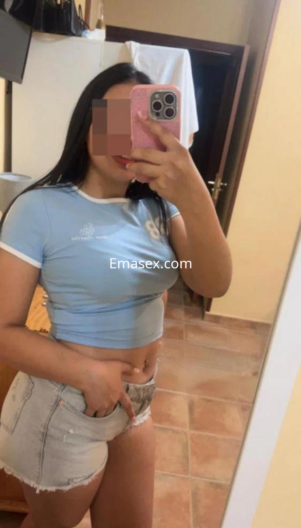 La puta en Toledo se muestra en un espejo, con una blusa azul y shorts cortos, sonriendo con confianza.
