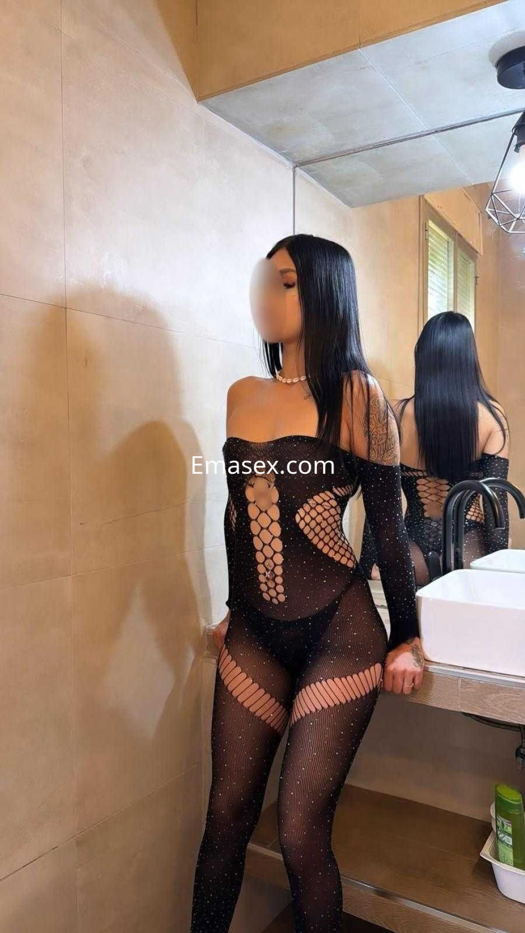 Escort en Toledo luciendo un body negro con detalles de malla, posando en un baño moderno con espejo.
