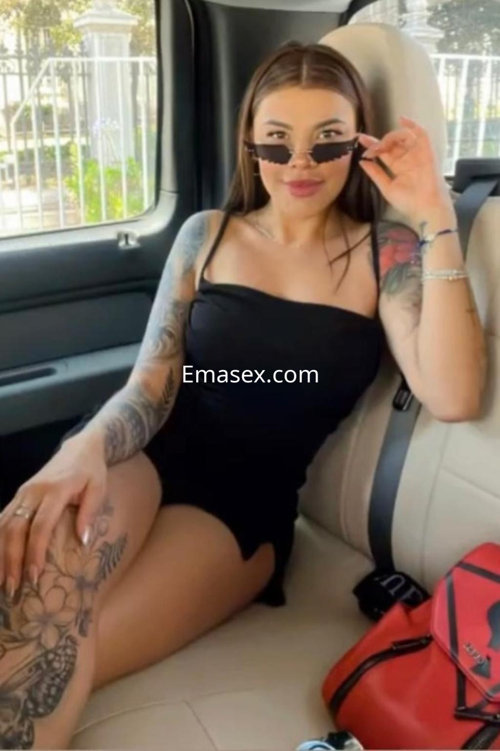 Puta sentada en un coche, luciendo un vestido negro ajustado y gafas de sol, con tatuajes visibles en sus brazos, en Girona.