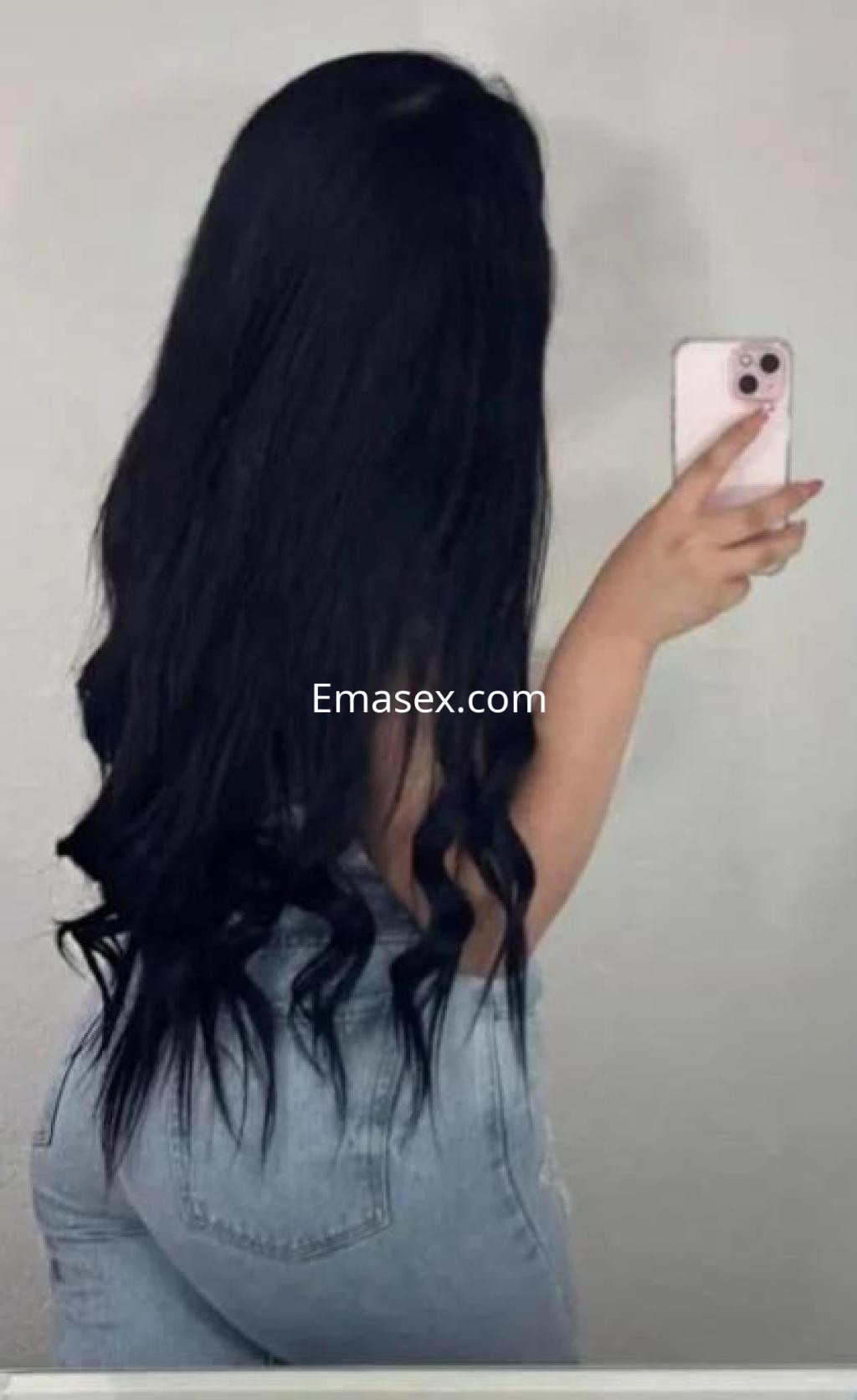 Puta de Lleida con largo cabello negro y ondulado, viste jeans ajustados y posa de espaldas frente al espejo.