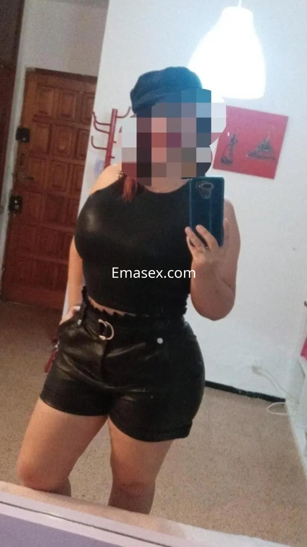 Escort en Oviedo con blusa negra ajustada y shorts de cuero, posando frente a un espejo en un entorno interior.