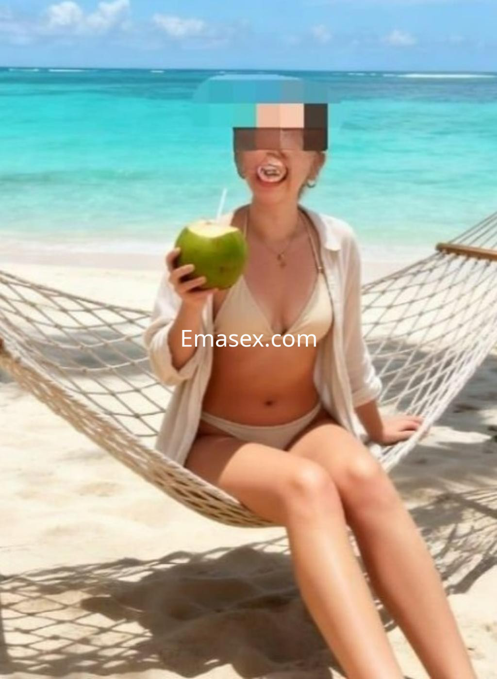Escort en Madrid, sentada en una hamaca en la playa, luciendo un bikini claro y sosteniendo un coco.