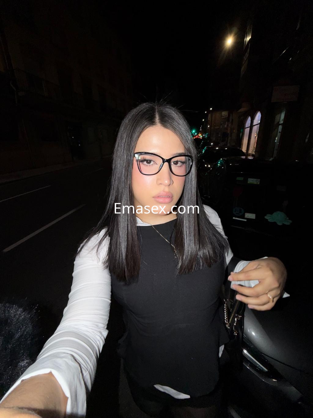 Escort trans en Vitoria, con cabello lacio y oscuro, luce una blusa blanca y gafas en un ambiente nocturno.