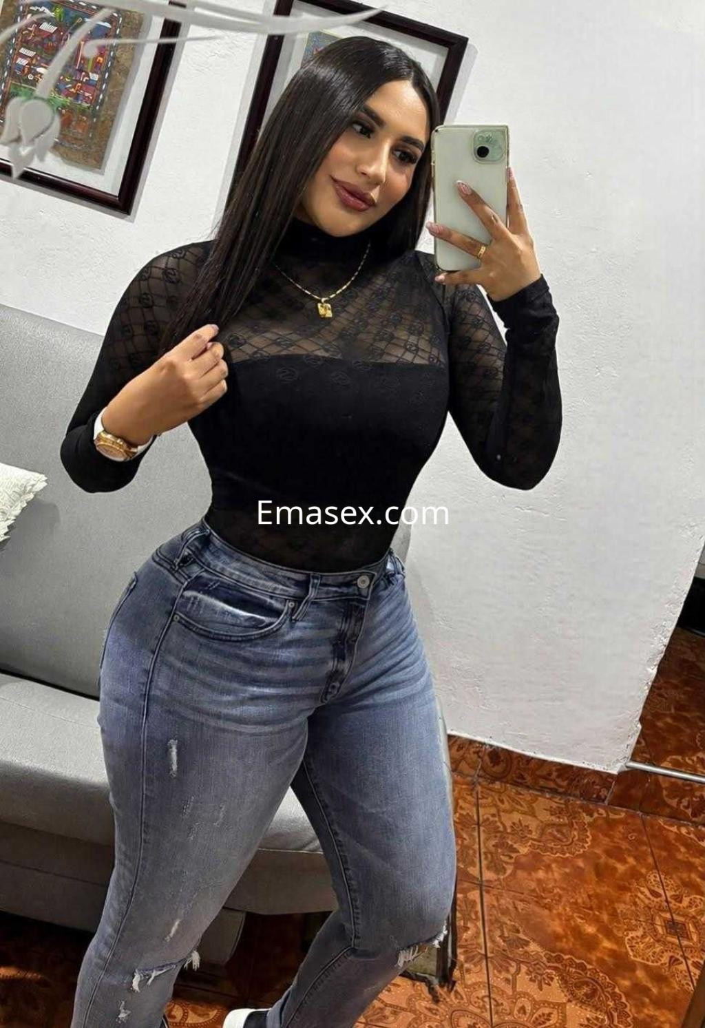 Puta en Alicante posa con un top negro transparente y jeans ajustados, frente a un espejo en un ambiente interior.