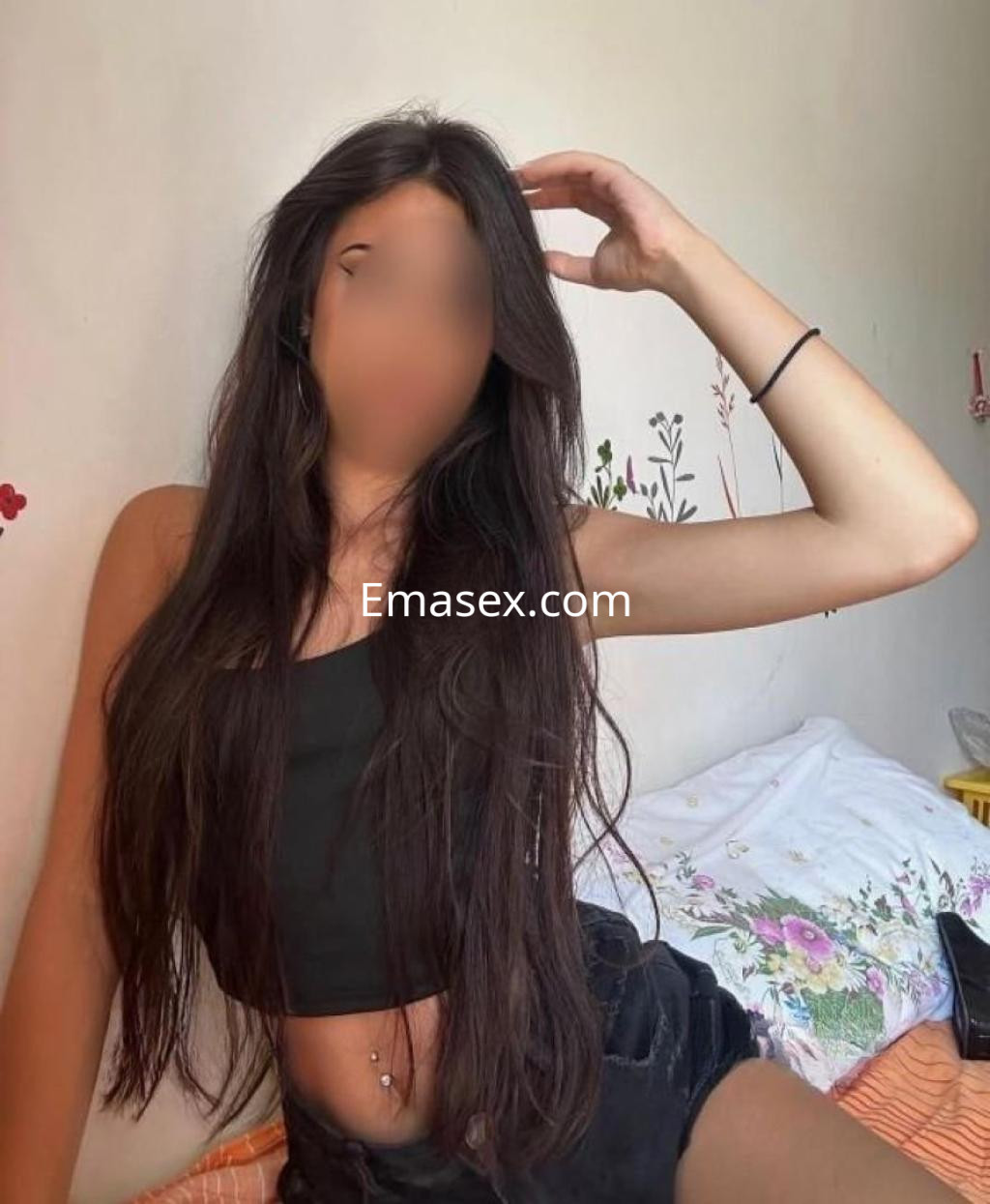 Escort con cabello largo y oscuro, usando un top negro y shorts, posando en una habitación con decoración floral en Almería.