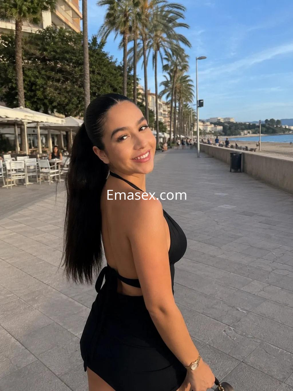 Puta posando en la playa de Palma de Mallorca, con un atuendo negro y una sonrisa radiante.