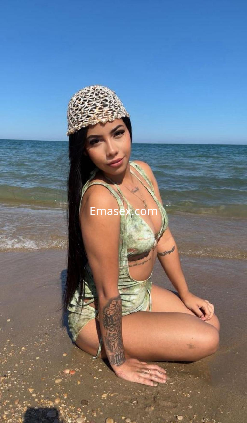 La escort se encuentra en la playa de Castellón, sentada sobre la arena con un traje de baño verde y un sombrero de tejido.