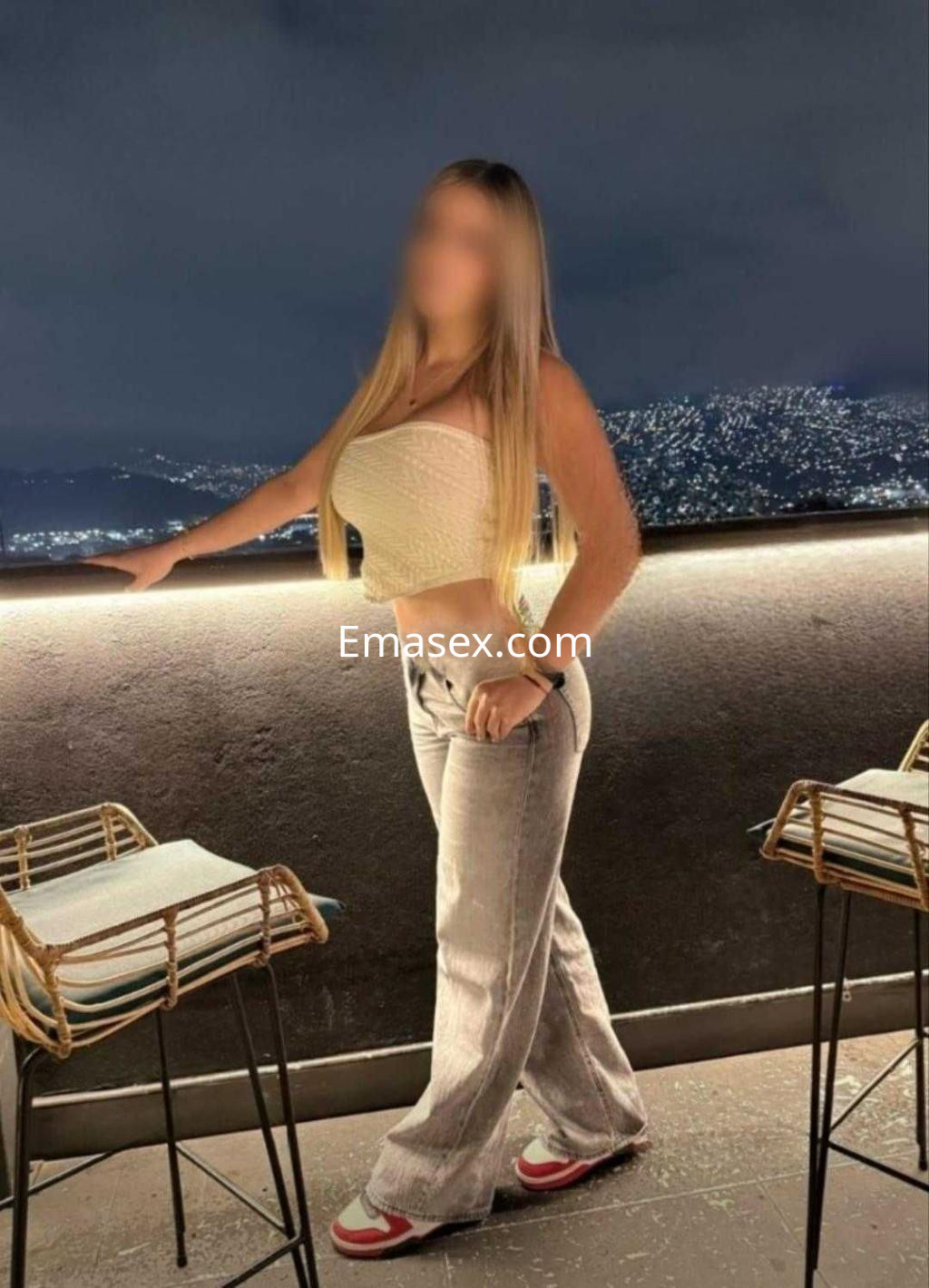 La puta luce un top ajustado y pantalones claros, posando en una terraza con vistas a Granada de noche.