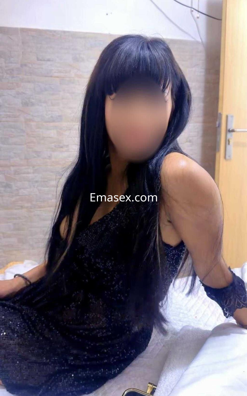 Puta en Granada con cabello largo y oscuro, vestida con un vestido negro brillante, sentada en una cama.