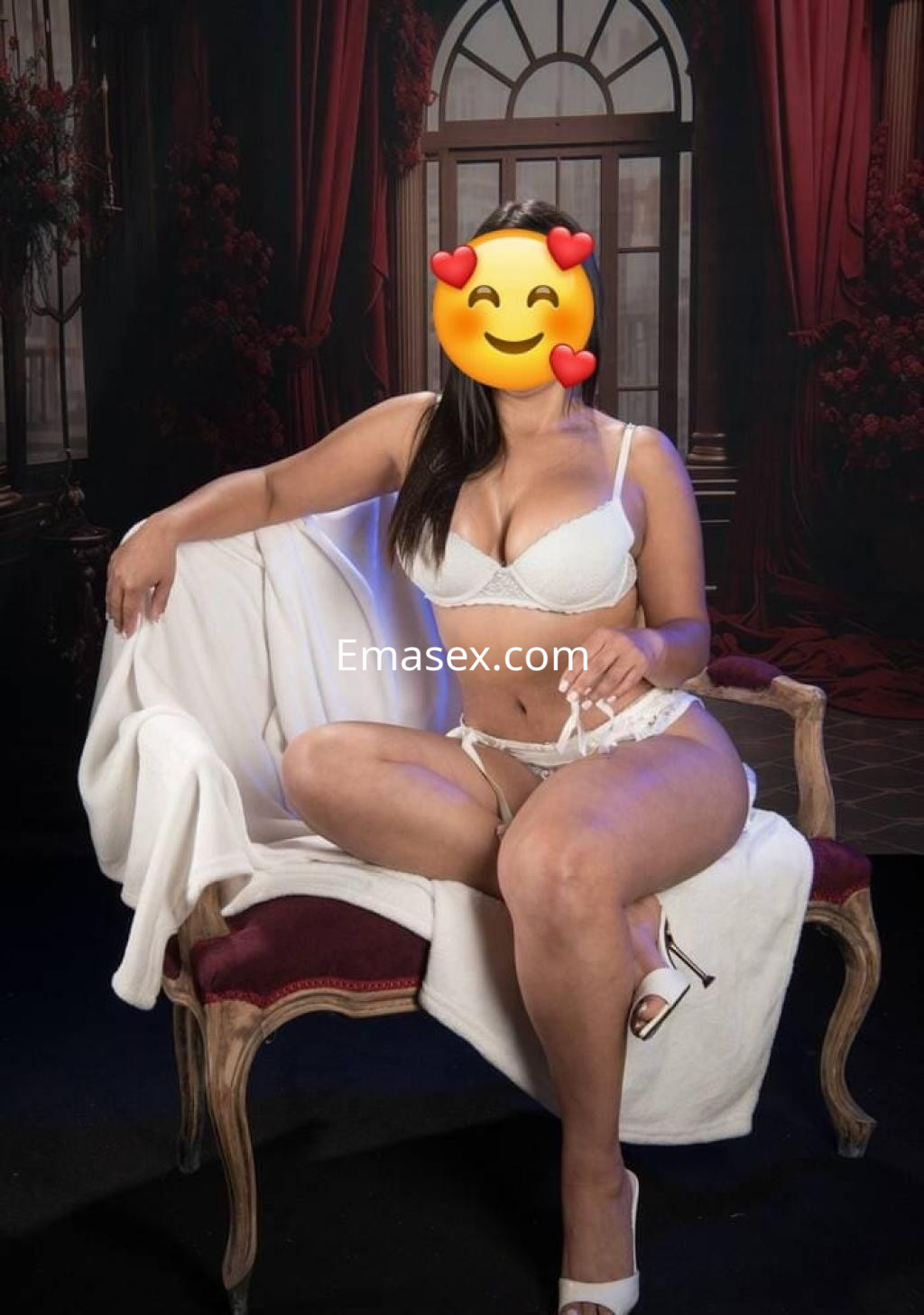 Escort en León, sentada en un sofá, viste lencería blanca y tacones altos, con fondo elegante.