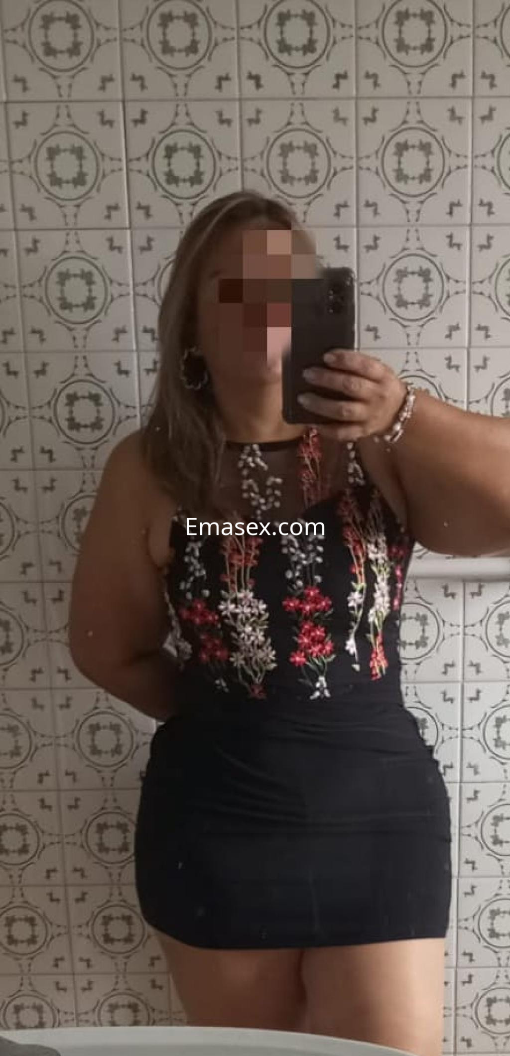 La puta posa frente al espejo con un vestido negro ajustado y detalles florales, en un entorno de Lugo.