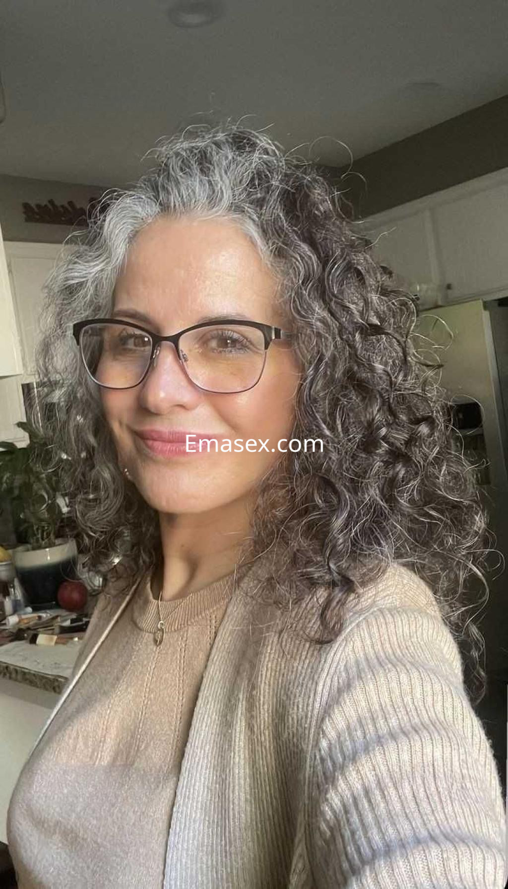 Escort con cabello rizado y canoso, usa gafas y un suéter claro, posando sonriente en un entorno acogedor en Madrid.