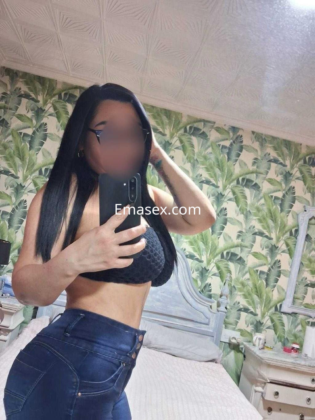 Puta en Murcia posando frente al espejo con blusa negra y jeans ajustados en un ambiente con decoración de plantas.